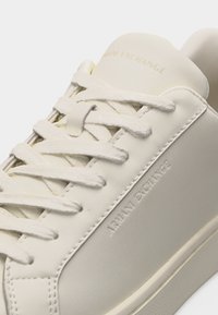 Αθλητικό παπούτσι Armani Exchange σε εκρού χρώμα με ταιριαστά κορδόνια και ανάγλυφο όνομα της μάρκας στο πλάι και στη γλώσσα, κοντινή όψη.