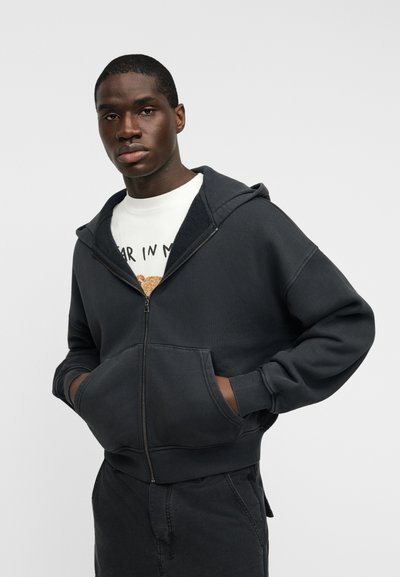 Μαύρο φούτερ με φερμουάρ, κουκούλα, μπροστινές τσέπες και άνετη εφαρμογή. Συνδυάζεται με λευκό κοντομάνικο μπλουζάκι με σχέδιο από κάτω. Λείο ύφασμα.