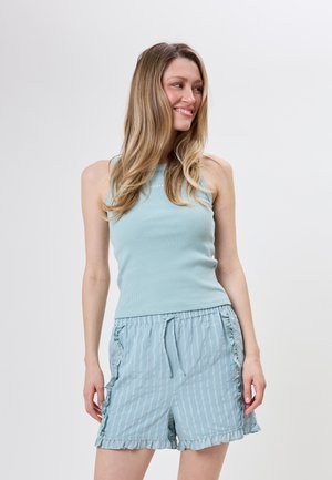 Ung kvinde smilende, iført en ærmeløs ribbet tanktop og lyseblå stribede shorts med flæsekant, står mod en ensfarvet baggrund.