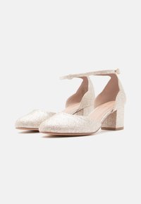 Trblietavé zlaté topánky na blokovom podpätku s špicatou špičkou, členkovým remienkom a mäkkou vnútornou podšívkou. Textúrovaný povrch odráža svetlo.