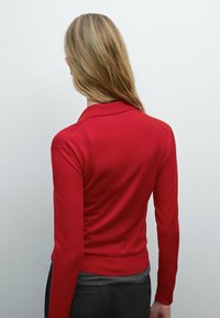 Blond persoon met lang haar draagt een rode lange mouw top en donkere broek, met de rug naar de camera gekeerd tegen een lichtgrijze achtergrond.