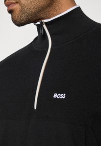 Μαύρος πουλόβερ με ψηλό γιακά και μισό φερμουάρ. Διαθέτει υφαμένο σχέδιο και λευκό λογότυπο "BOSS" στο στήθος.