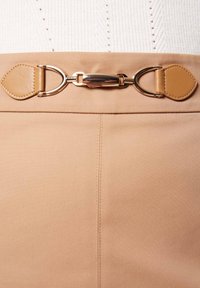 Jupe couleur fauve avec une finition lisse et une boucle en métal doré ornée de détails en cuir à la taille. Les coutures sont visibles.