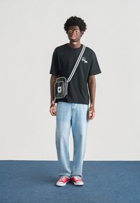 Μαύρο t-shirt με γραφικό λογότυπο, φαρδιά ανοιχτό μπλε τζιν, κόκκινα αθλητικά παπούτσια και μια μικρή μαύρη τσάντα χιαστί με λευκά στοιχεία.