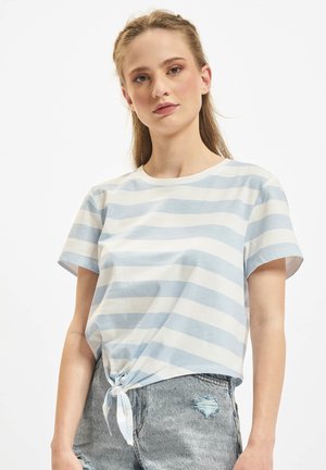 Junge Frau mit hellbraunem geflochtenem Haar, die ein blau-weiß gestreiftes kurzärmliges Shirt trägt, das um die Taille gebunden ist, sowie zerrissene Denim-Shorts.