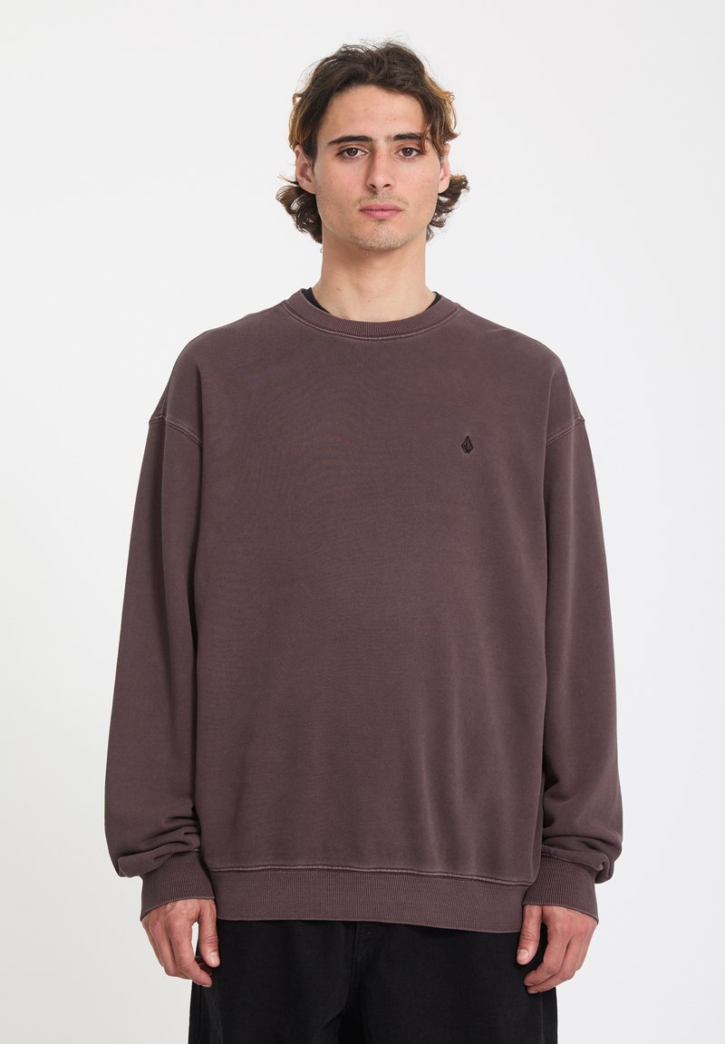 Jeune homme aux cheveux mi-longs portant un sweat-shirt à col rond ample et délavé de couleur violette et un pantalon noir, debout devant un fond blanc.
