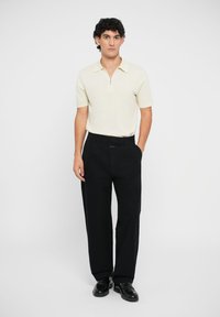 Polo de manga corta en color crema con cuello de cremallera, combinado con pantalones negros y zapatos negros, con un corte recto y una textura suave.