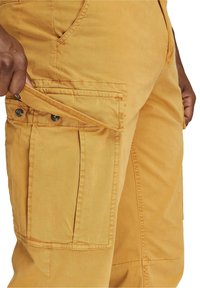 Cargo byxor i senapsgul tyg med flera fickor, med tryckknappsstängningar och synliga söm detaljer. Texturerad yta.