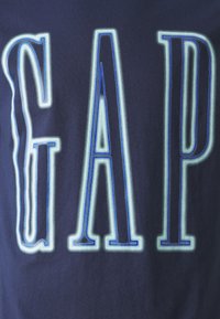 Tmavě modrá tkanina s velkým, obrysovým nápisem "GAP" napsaným velkými písmeny, který září bílým a modrým neonovým efektem.