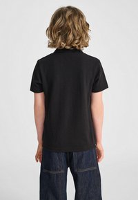 Barn med axellångt lockigt hår som bär en enfärgad svart kortärmad tröja och mörkblå jeans, vänd bort från kameran mot en vit bakgrund.