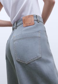 Lys grå højtaljede jeans med et lædermærke med præget design på bagsiden, synlig søm og dobbelte baglommer.