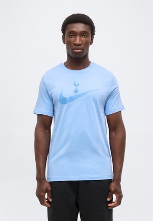 Homme portant un t-shirt bleu clair avec un grand swoosh Nike et un logo coq au-dessus, associé à un pantalon foncé, sur un fond uni.