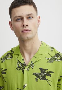Camisa verde de manga corta con patrones florales negros, con cuello y botones claros, hecha de una tela suave.