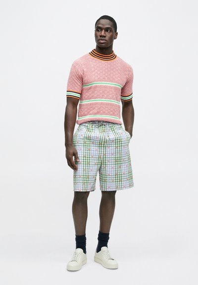 Κόκκινο πλεκτό πουλόβερ με κοντά μανίκια και ριγέ γιακά, συνδυασμένο με πράσινα καρό σορτς με λουλουδένια σχέδια. Στο πόδι λευκά αθλητικά παπούτσια.