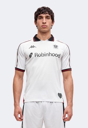 Jeune homme portant un maillot de football blanc avec des bordures noires et rouges, logo du sponsor Robinhood et un emblème en forme de cœur près de l'ourlet.