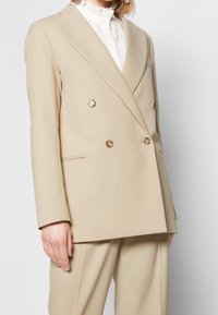Frau in einem beigen, doppelt geknöpften Blazer mit passender Hose und einer weißen, rüschengeschmückten Bluse mit hohem Kragen.