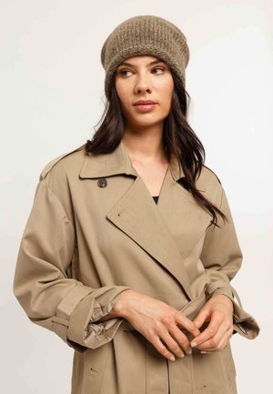 Beige trench coat with oversized sleeves and a wide collar, worn over a black top. Brown ribbed beanie adds texture to the outfit.