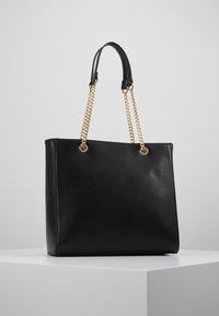 Czarna skórzana torba typu tote z uchwytami z złotego łańcucha i czarnymi skórzanymi uchwytami na ramię, wyeksponowana na białym cokole.