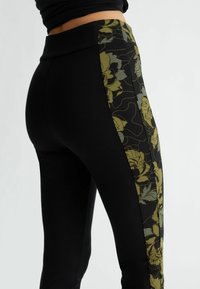Leggings negros con un ancho panel lateral estampado con diseños florales en tonos verdes y apagados. Textura suave y diseño ajustado.