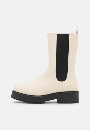 Flødefarvet støvle til midt på læggen med sort elastisk sidepanel, sort kraftig sål, rund tå og gul trækstrop bagpå.