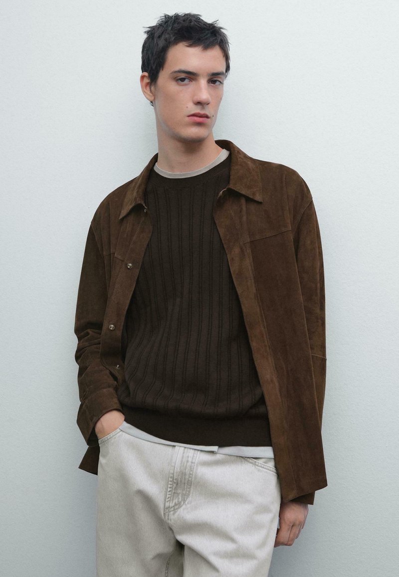 Jonge man die tegen een eenvoudige muur staat, gekleed in een bruine suede jasje, een donkerbruine trui en lichtbeige broek, met één hand in zijn zak.