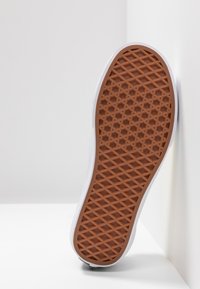 Sole of a white sneaker resting against a plain wall, highlighting its textured brown rubber bottom with a diamond-pattern design.