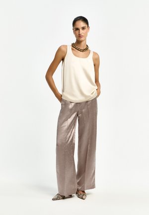 Frau trägt ein cremefarbenes ärmelloses Top, metallische Hosen mit weitem Bein, spitze Schuhe mit Schlangenmuster und eine auffällige goldene Halskette, steht mit den Händen in den Taschen.