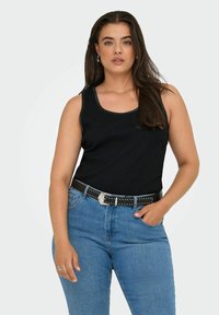 Vrouw met lang donker haar, gekleed in een zwarte mouwloze top, blauwe spijkerbroek en een zwarte riem met studs, staand met één hand in de zak.