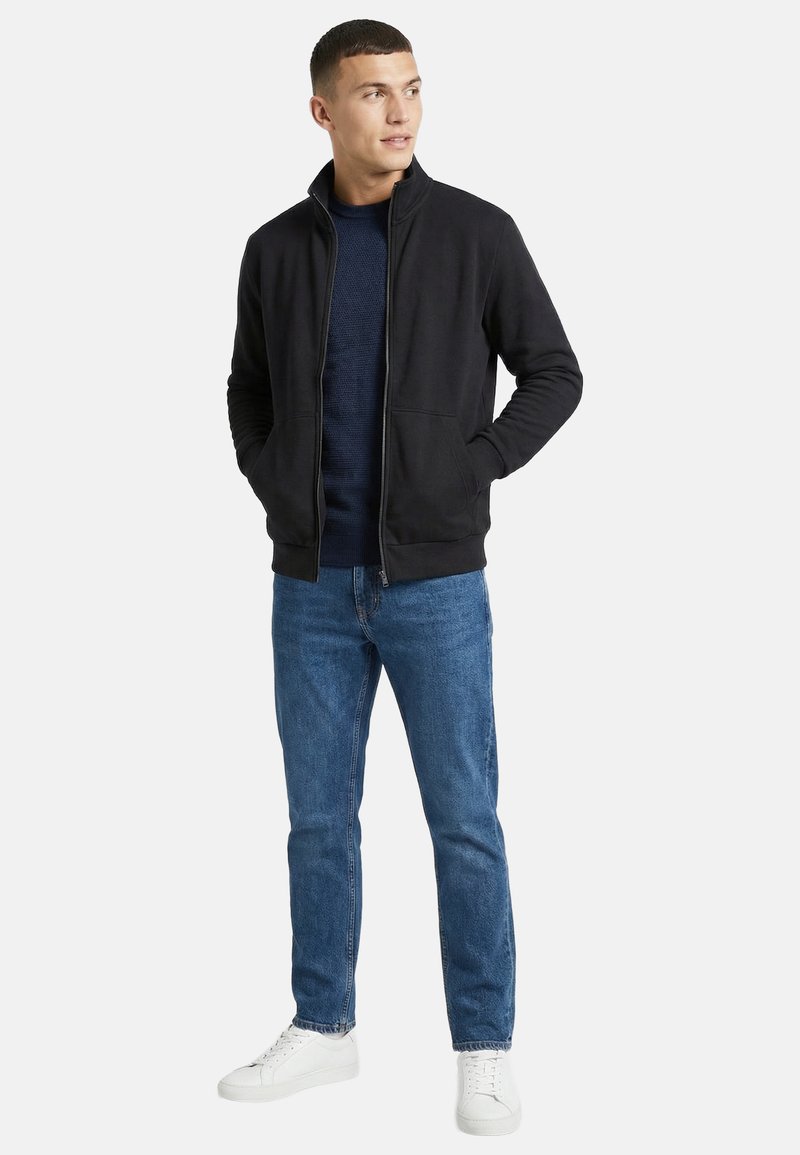 Homme debout, les mains dans les poches, portant une veste noire zippée, un pull bleu marine, un jean bleu et des baskets blanches, regardant vers la droite.