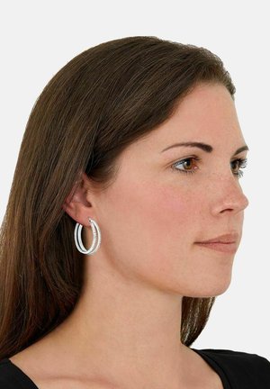 Pendientes de aro plateados con un diseño circular en capas, que presentan un acabado texturizado y bordes lisos, llevados por un modelo con cabello largo castaño.