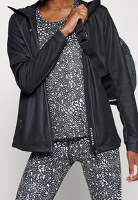 Giacca da atletica nera con una texture liscia, tasche con zip, abbinata a una maglietta a pois bianco e nero e leggings con un motivo simile.