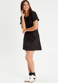 Robe noire à manches courtes avec une finition texturée, une taille ceinturée et une coupe décontractée, portée avec des baskets blanches et des chaussettes noires à motifs.