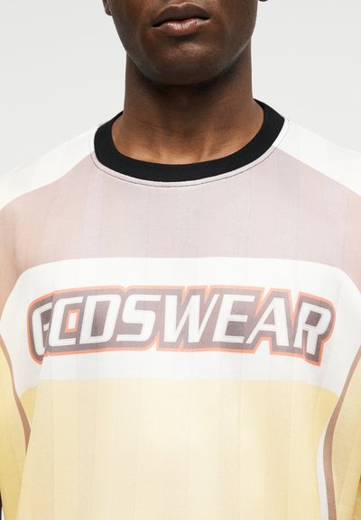 Άνδρας που φοράει μπλουζάκι με μπλοκ χρώματος σε μπεζ, λευκή και κίτρινη περιοχή με το κείμενο «GCDSWEAR» στο στήθος.