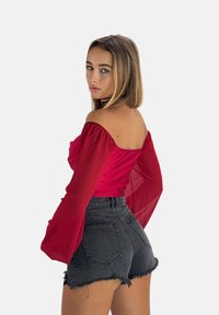 Haut rouge décolleté en satin avec des manches transparentes, associé à un short taille haute noir déchiré. Fond épuré.