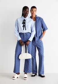Două modele stau lângă un perete alb; amândouă poartă pantaloni texturați de culoare albastră. Una poartă o cămașă albastră deschisă cu accente negre din panglică, ținând o geantă albă.