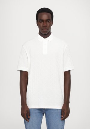 Camisa polo blanca de manga corta con un patrón texturizado, que cuenta con un cuello y una botonera, combinada con jeans azules.