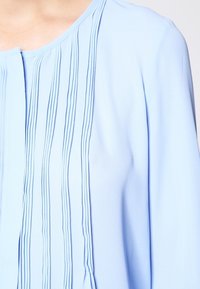 Blusa azzurra chiara con pieghe verticali, scollo rotondo e maniche lunghe, realizzata in tessuto morbido e leggero con una texture liscia.