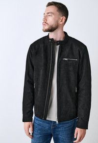 Sort suedeskindjakke med stående krave, to sidelommer og en brystlomme med lynlås, prydet med sølvhardware og en glat overflade.