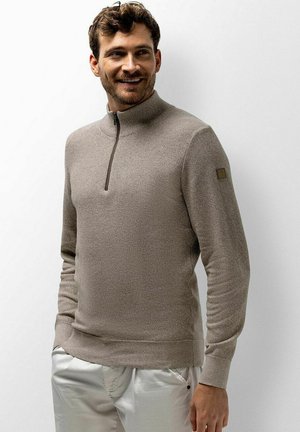 Mann in beigem Pullover mit Viertelreißverschluss und weißen Hosen steht lächelnd mit den Händen in den Taschen vor hellem, einfachem Hintergrund.
