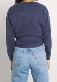 Femme portant un pull en maille bleu marine et un jean taille haute bleu clair avec des poches arrière, se tenant devant un fond blanc.