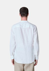 Camicia bianca di lino a maniche lunghe con orlo arrotondato, colletto casual e polsini con bottoni. Il tessuto appare texture e leggero.