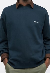 Homme portant un sweat-shirt bleu marine avec le texte blanc « NN. 07 » sur la poitrine, superposé à une chemise marron à col, mains dans les poches d’un pantalon kaki.