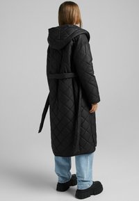 Czarny pikowany długi płaszcz z kapturem i paskiem, charakteryzujący się gładką fakturą, dużą osłoną i bocznymi kieszeniami. Nosi się go na jasnoniebieskie dżinsy i masywne czarne botki.