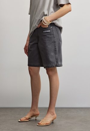 Person iført mellemgrå mørkegrå shorts med "PEGADOR" logo, en løs lysegrå t-shirt, cremefarvede højhælede sandaler og lagdelte armbånd.