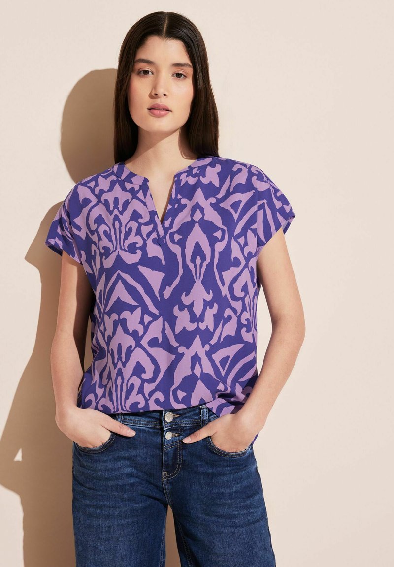 Camicetta viola a maniche corte con scollatura a V, caratterizzata da un motivo floreale in tonalità più chiare. Indossata con jeans in denim blu.