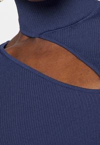 Marinblå ribbad topp med hög krage, utskärningsdesign på axlarna och slät yta som betonar unik form och detaljer.