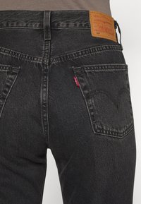 Jeans de mezclilla negra con un diseño clásico, que presentan un parche de cuero en la cintura y una etiqueta roja en el bolsillo trasero.