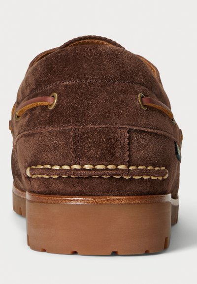 Zapato náutico de ante marrón con cordones de cuero atravesados por ojales metálicos y una suela de goma gruesa, visto desde atrás.