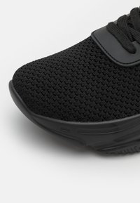 Chaussure de sport noire avec une tige en mesh texturé et une semelle en caoutchouc, présentant un design à lacets et un bout plat et arrondi.