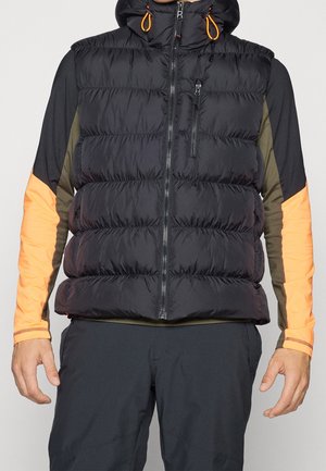 Homme portant une veste matelassée noire avec capuche par-dessus une chemise à manches longues avec des manches orange et vert olive, et un pantalon noir.